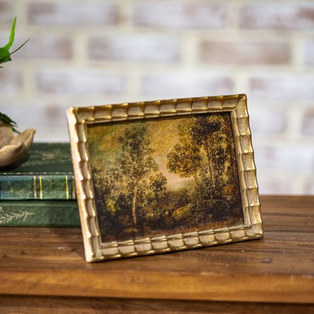Framed forest landscape painting in a scalloped gold frame on a wooden surface next to stacked books.