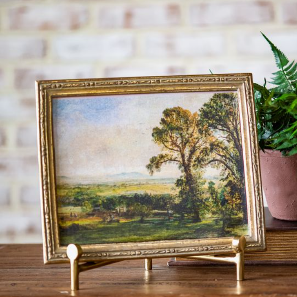 Large framed landscape painting with tall trees in a gold frame, displayed on a metal easel.