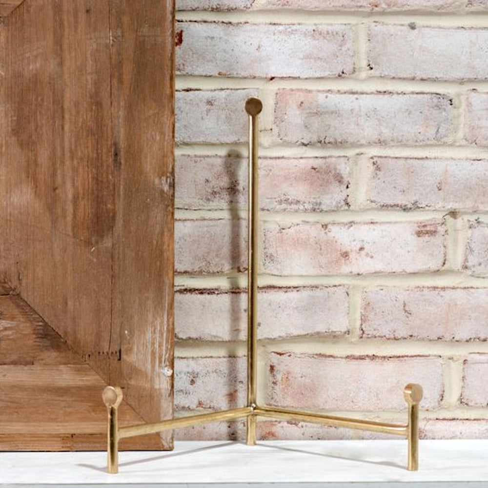 Large brass straight-back easel for displaying plates or art, set against a white brick wall and wooden panel.