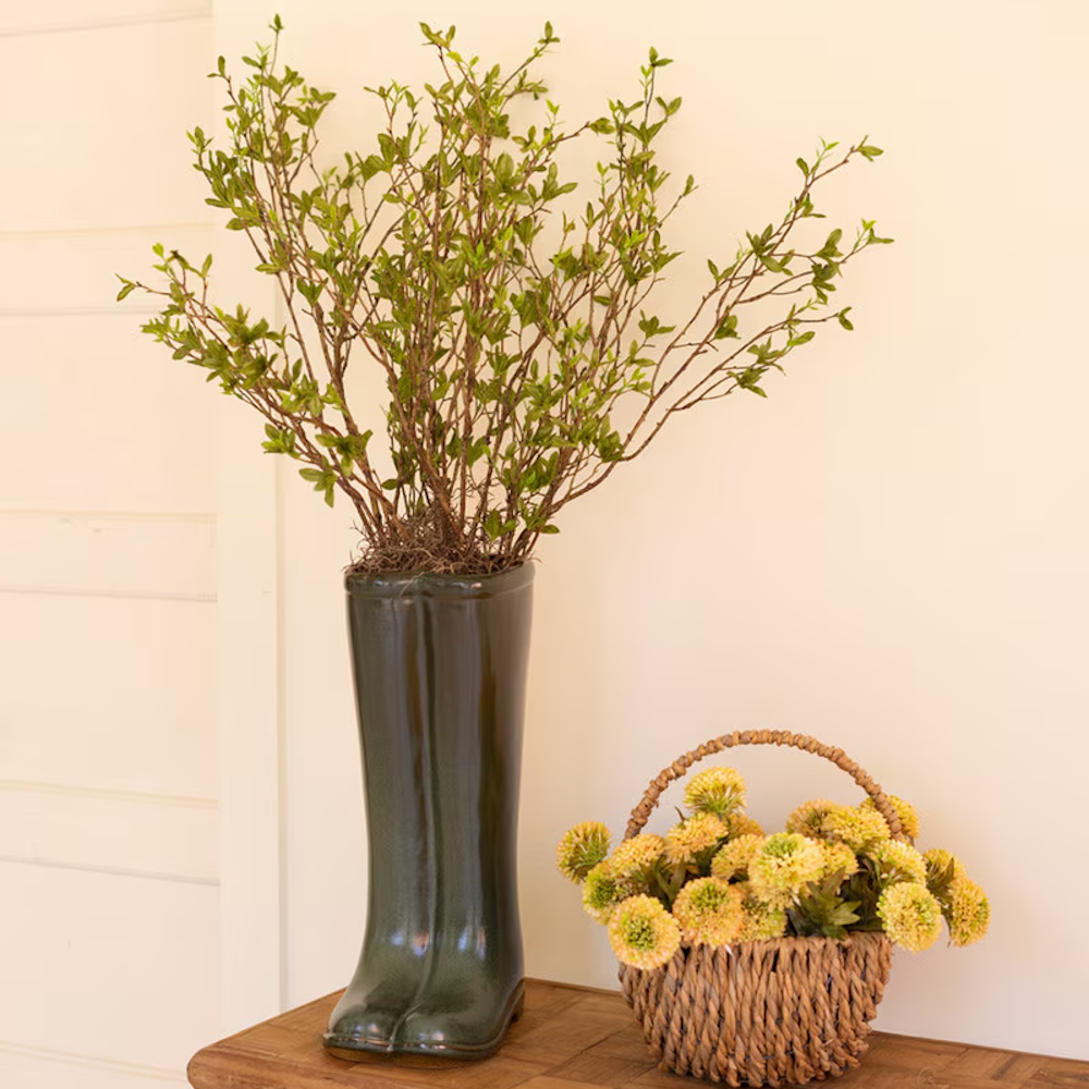 Green ceramic boot vase with leafy branches next to a woven basket of yellow flowers on a wooden table.