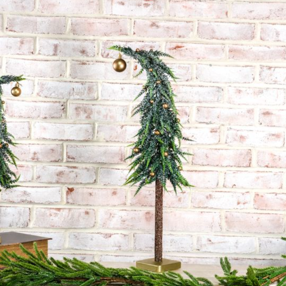 Frosted mini tabletop evergreen tree with gold ornaments on a stand, set before a white brick wall.