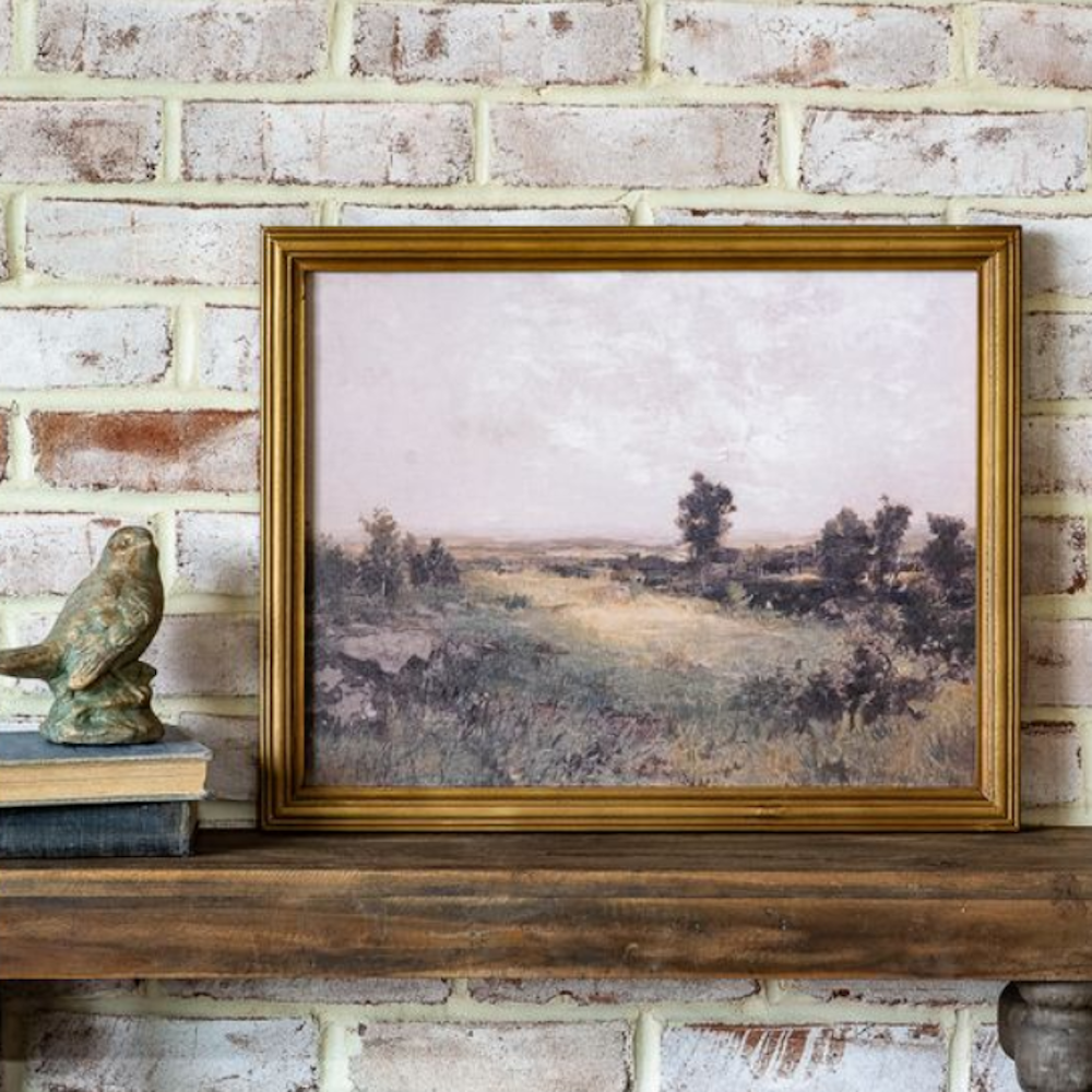 Framed open field landscape painting in a gold frame, sitting on a mantle next to a bird figurine.