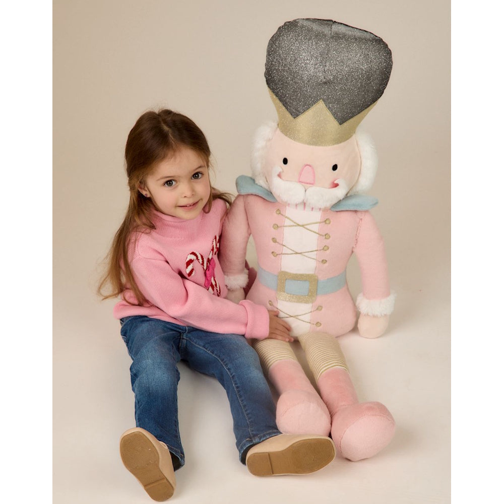 Little girl sitting next to a large pink plush nutcracker toy on a white studio background.