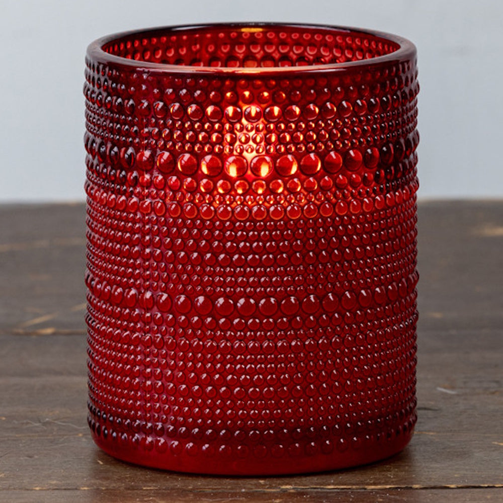 Lit candle inside a red beaded glass holder sitting on a dark wooden surface.