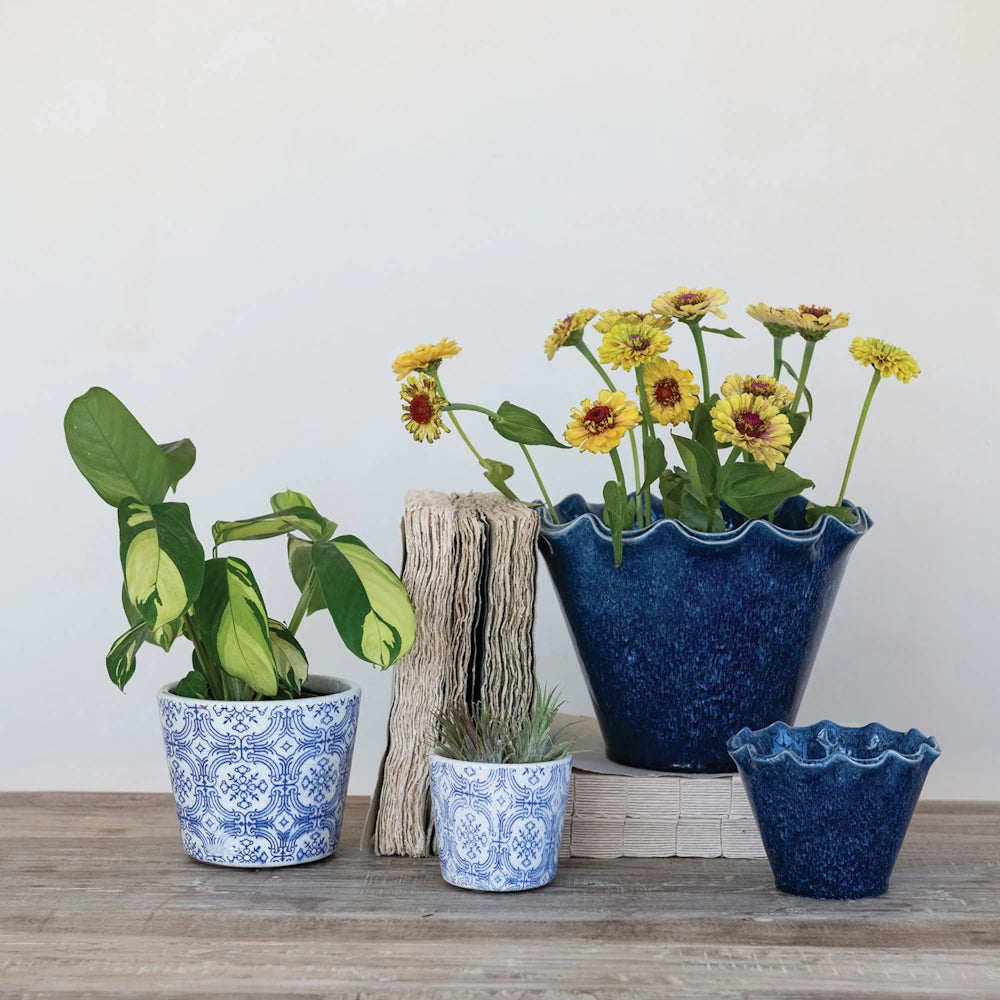 Blue and white patterned ceramic planters and ruffled stoneware pots with yellow flowers on a rustic wooden table.