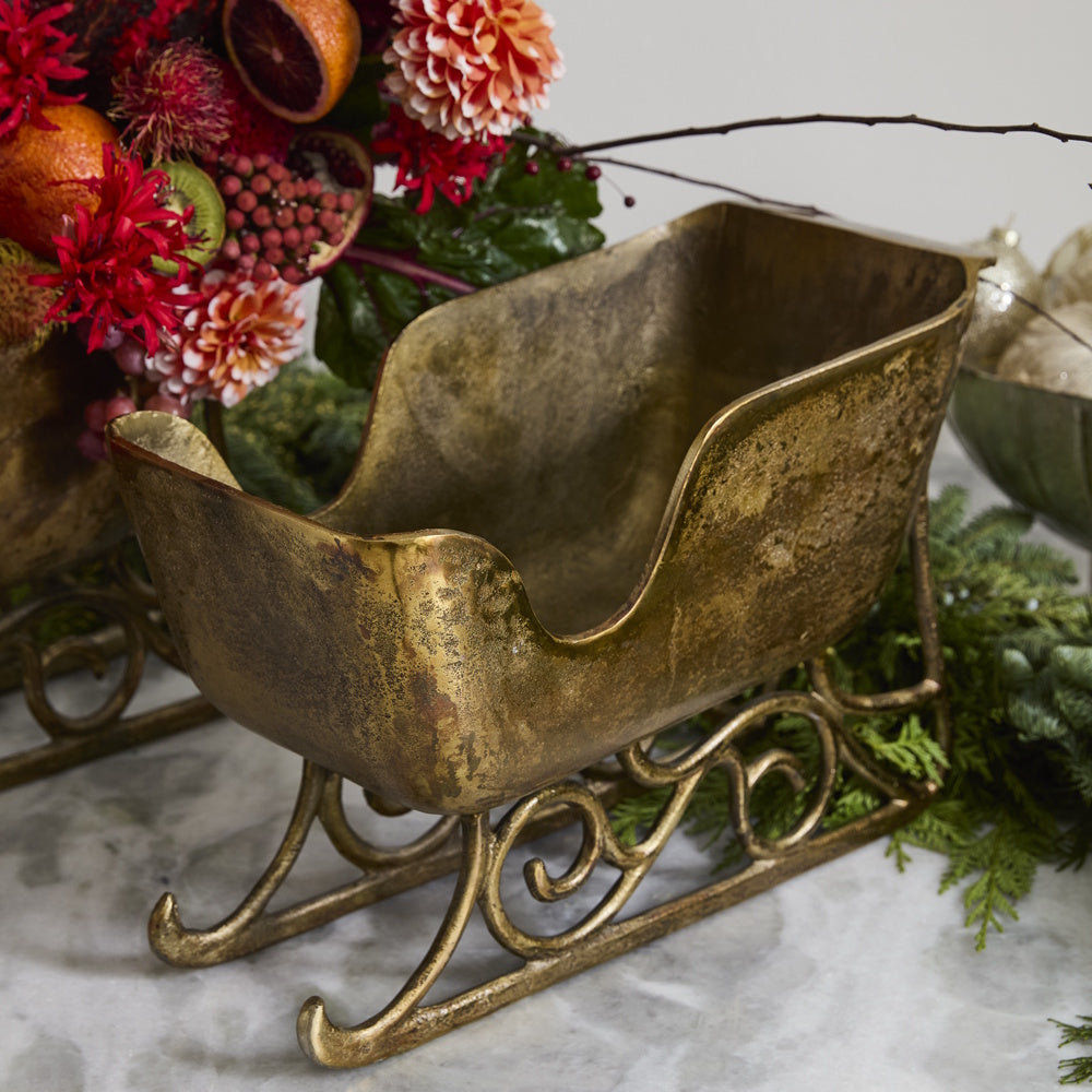 Vintage-style brass sleigh planter with decorative scroll metal runners on a marble surface.
