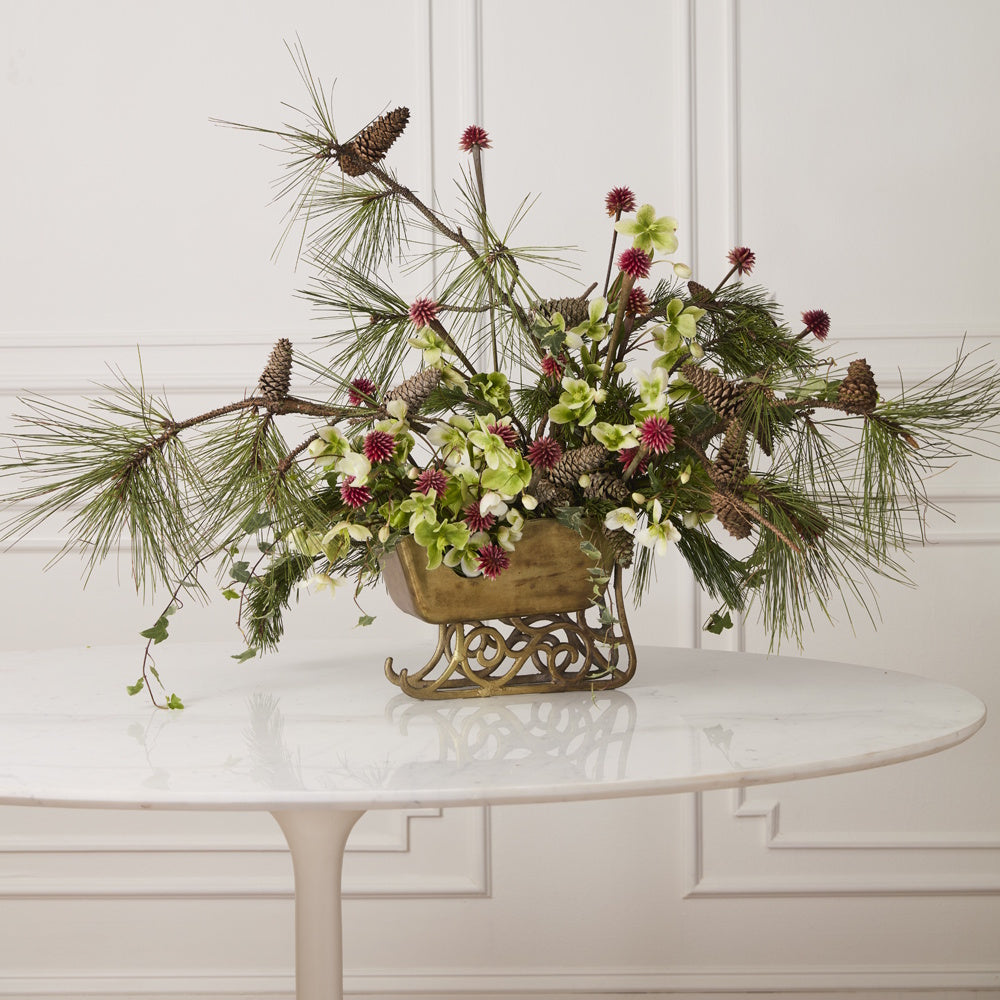 Brass sleigh planter filled with pine branches, pinecones, and flowers on a white table.