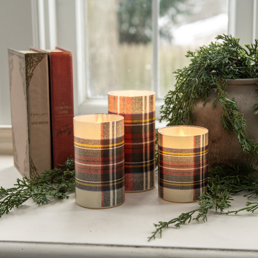 Three glowing flameless pillar candles wrapped in a white tartan plaid pattern by books and greenery.