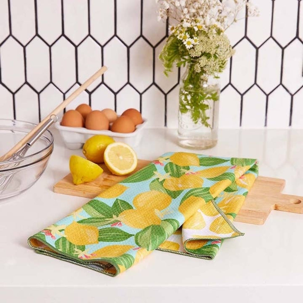 Lemon patterned tea towel on a wooden board with fresh lemons, eggs, and white flowers in a modern kitchen.