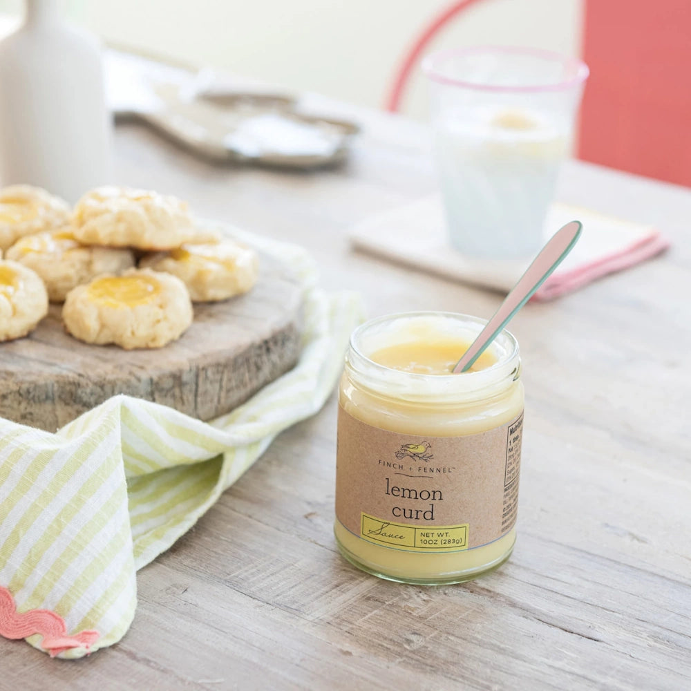 An open jar of Finch + Fennel lemon curd sauce with a spoon, next to a platter of lemon thumbprint cookies.
