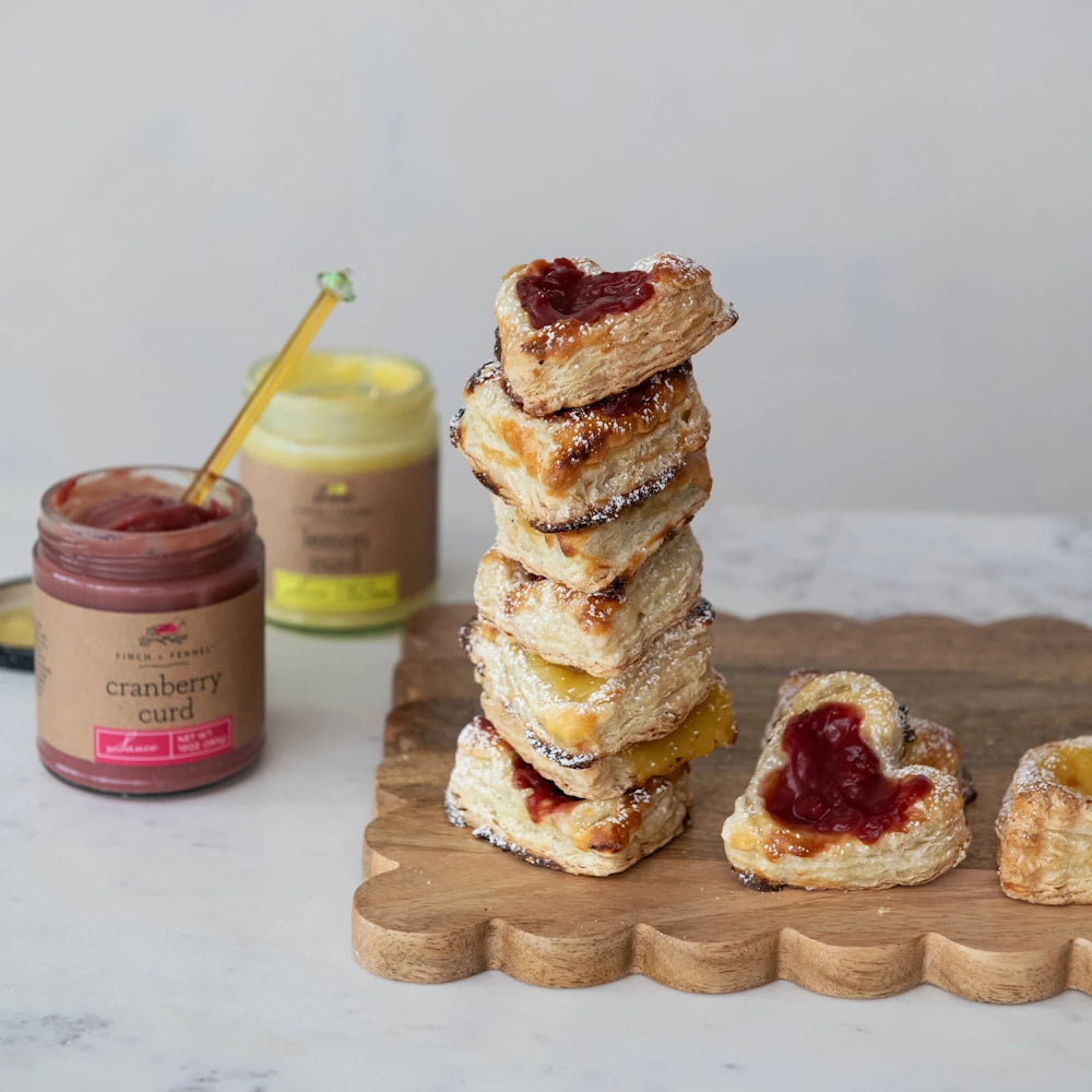 Stack of heart-shaped puff pastries filled with cranberry and lemon curd next to jars of Finch & Fennel sauce.