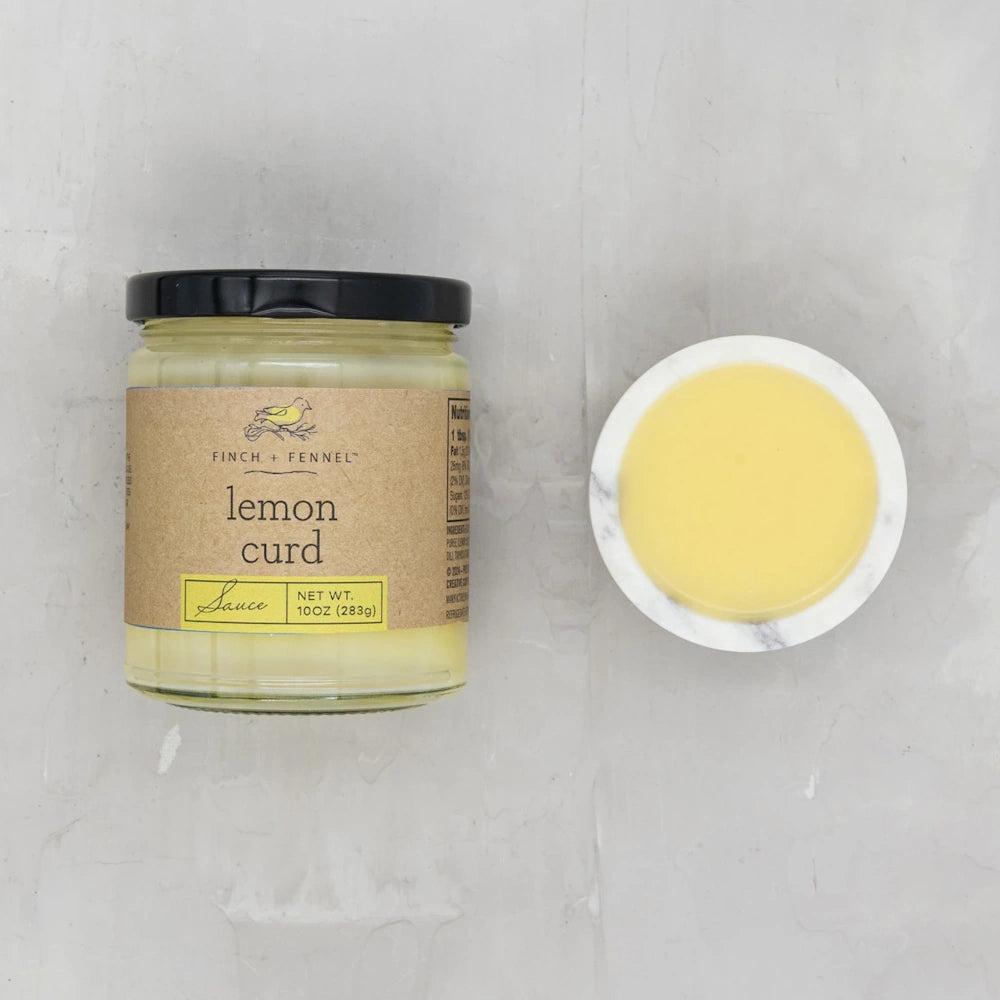 A glass jar of Finch + Fennel Lemon Curd Sauce next to a small marble bowl of the yellow sauce on a grey surface.