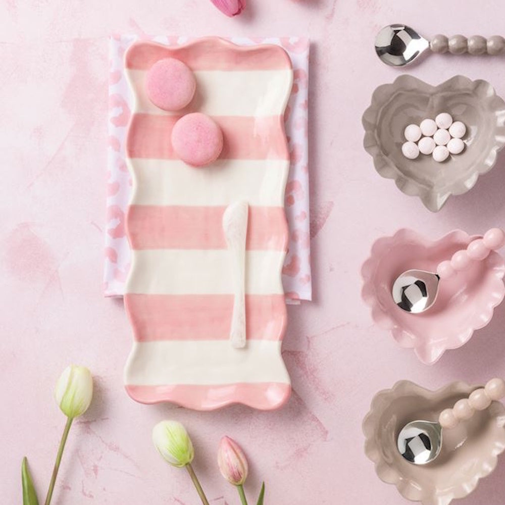 Pink and white striped ceramic tray with heart-shaped bowls, beaded spoons, and macarons on a pink background.