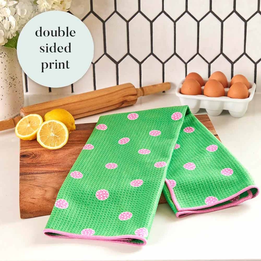 Green kitchen tea towel with pink pickleball pattern on a wooden board next to lemons, rolling pin, and eggs.