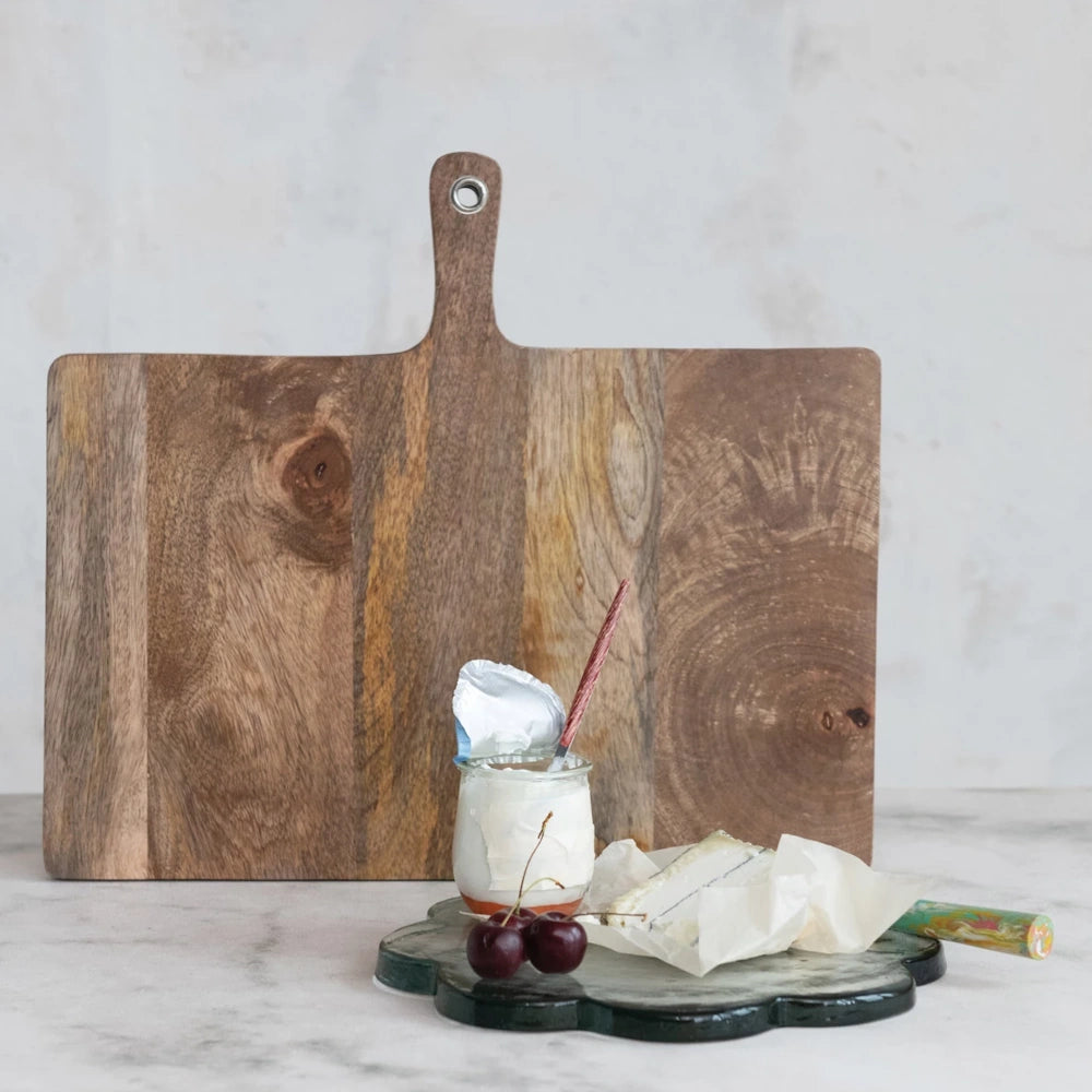 Wooden charcuterie board behind a green scalloped glass serving tray with brie cheese, cherries, and a yogurt jar.