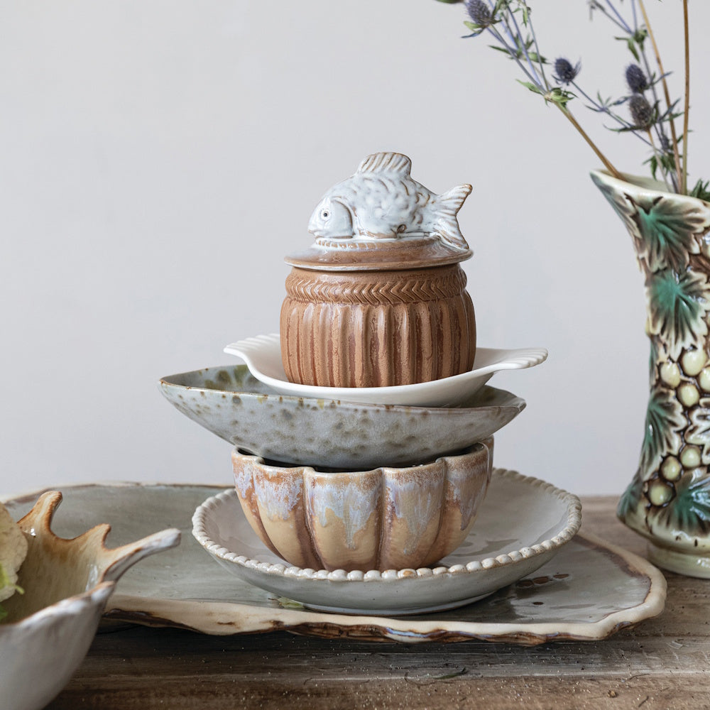 Rustic stoneware salt cellar with a decorative fish lid stacked on ceramic bowls and plates for a coastal table setting.