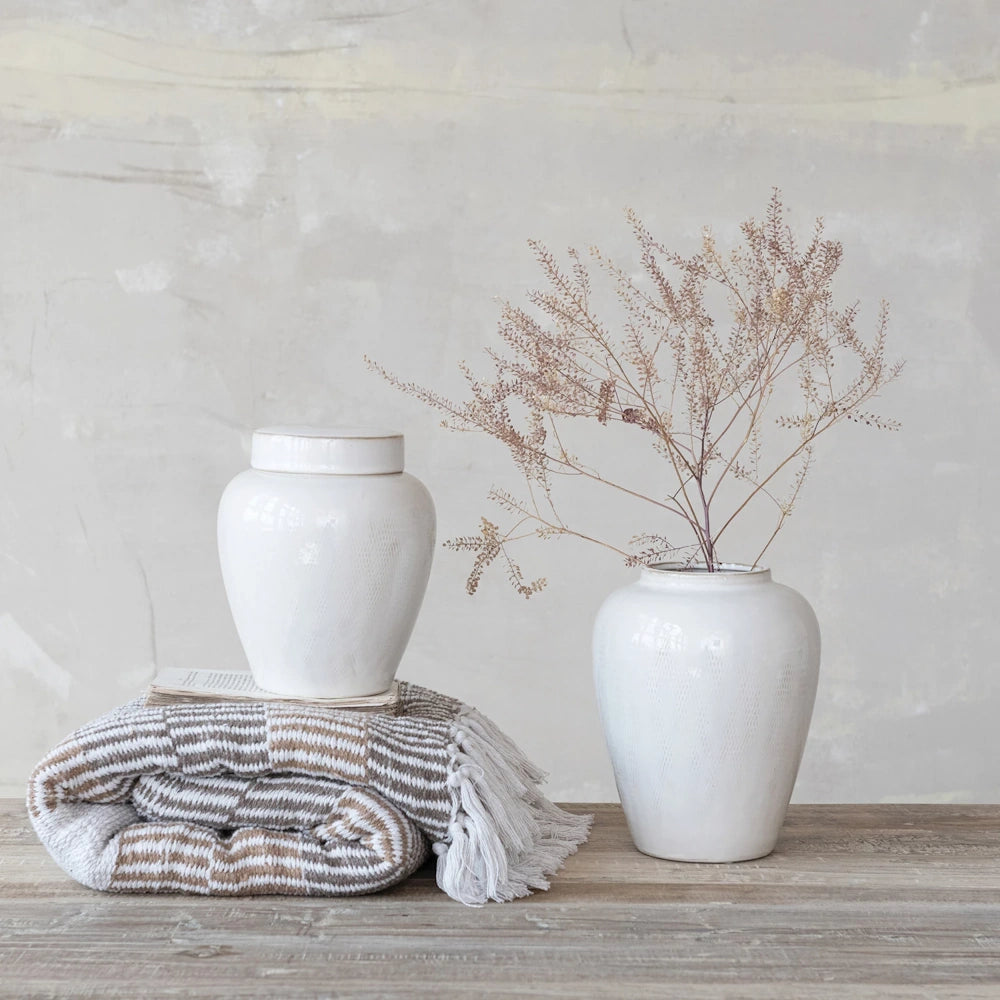 White ceramic ginger jars and a striped tassel throw blanket on a rustic wooden table with dried floral stems.