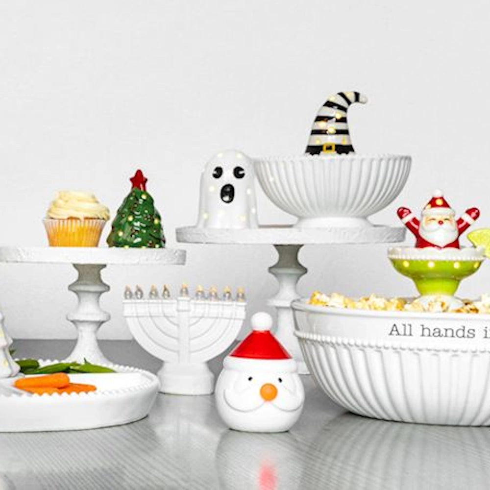 Holiday table display featuring ceramic light-up sitters, white cake stands, and serving bowls.