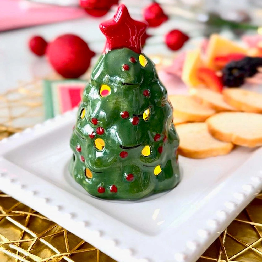 Lit ceramic Christmas tree sitter on a white serving platter next to snacks and holiday decor.