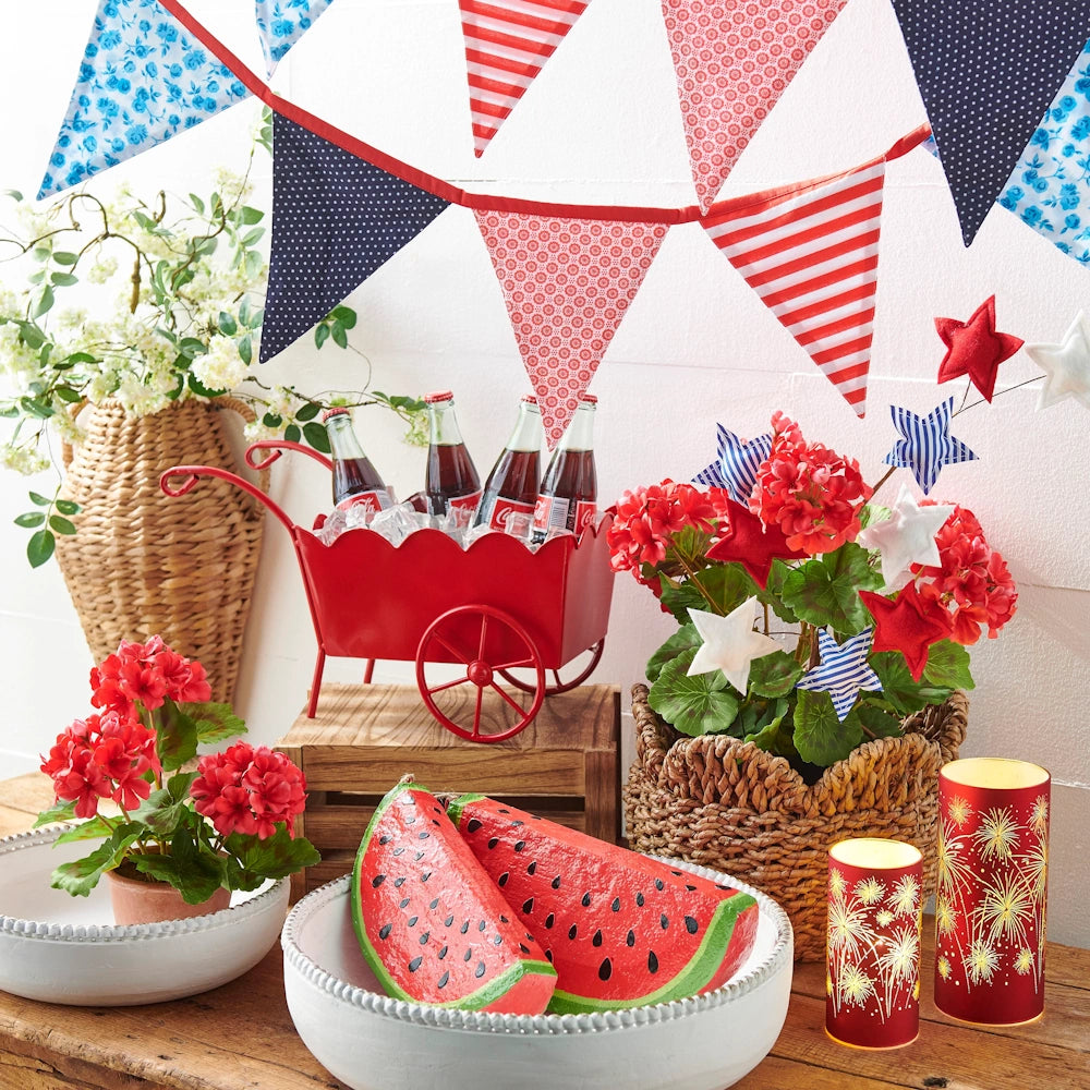 Festive 4th of July decor with red, white, and blue bunting, vibrant geraniums, star ornaments, watermelon slices, and bottles in a red wagon.