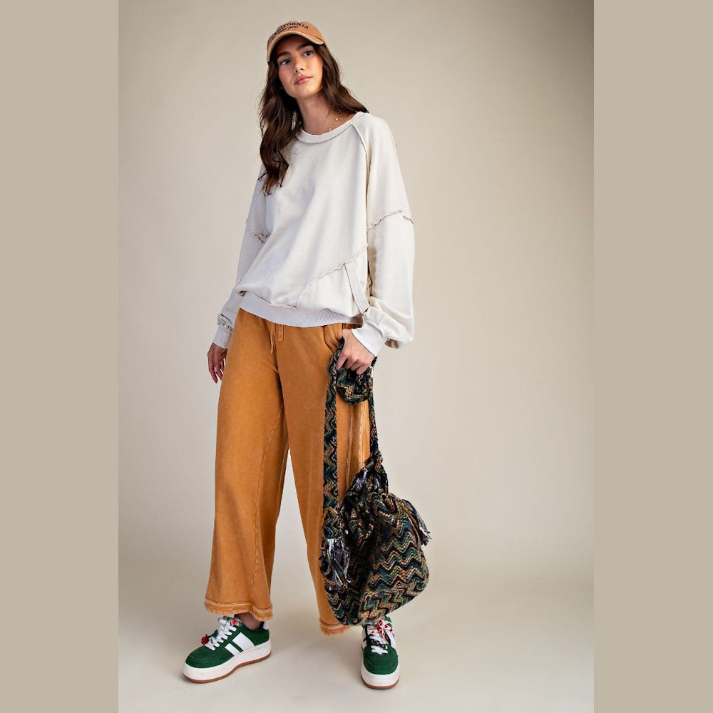 Woman in a cream long-sleeve mineral wash pullover, mustard wide-leg pants, and green sneakers with a patterned bag.