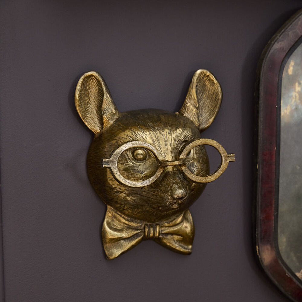 Gold wall-mounted mouse head sculpture wearing round glasses and a bow tie against a dark purple background.