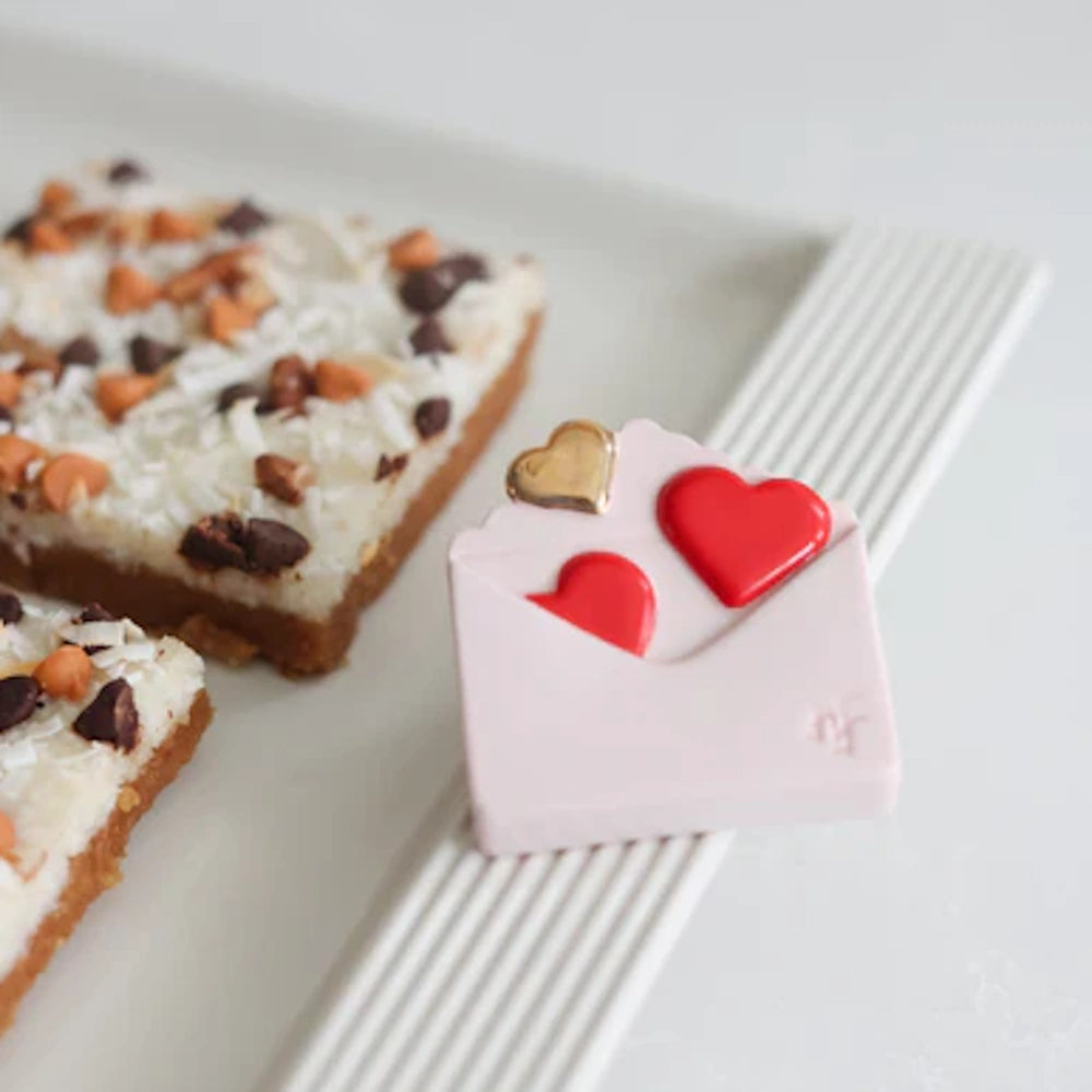 A pink envelope-shaped chocolate with red and gold hearts next to coconut and chocolate chip dessert bars on a white tray.