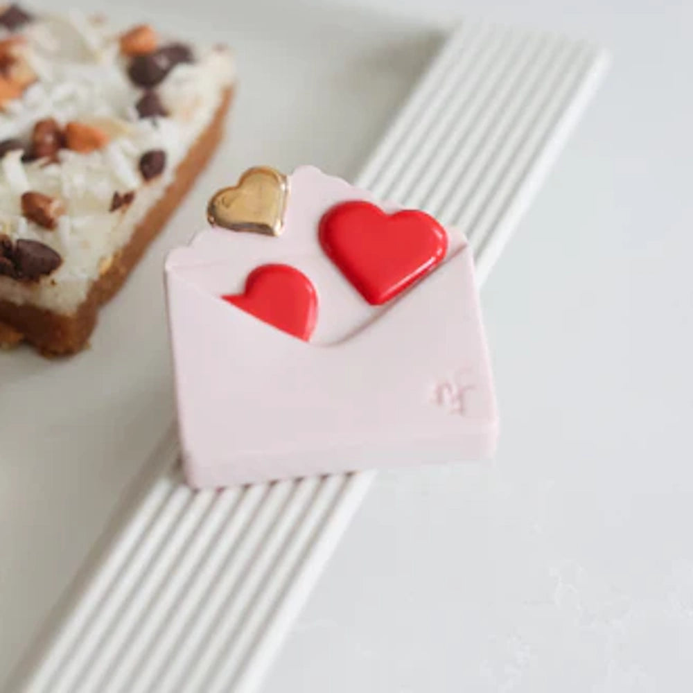 Pink envelope-shaped decorative item with red and gold hearts on a white ribbed tray next to a dessert bar.