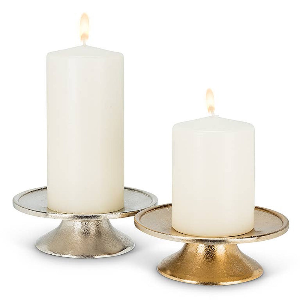 Two lit white pillar candles resting on silver and gold textured metal pedestal stands.