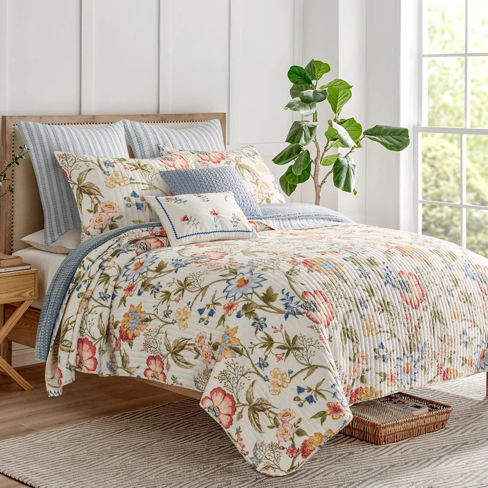 Bed styled with multicolor floral quilt, striped euro shams, decorative throw pillows, and a plant nearby.