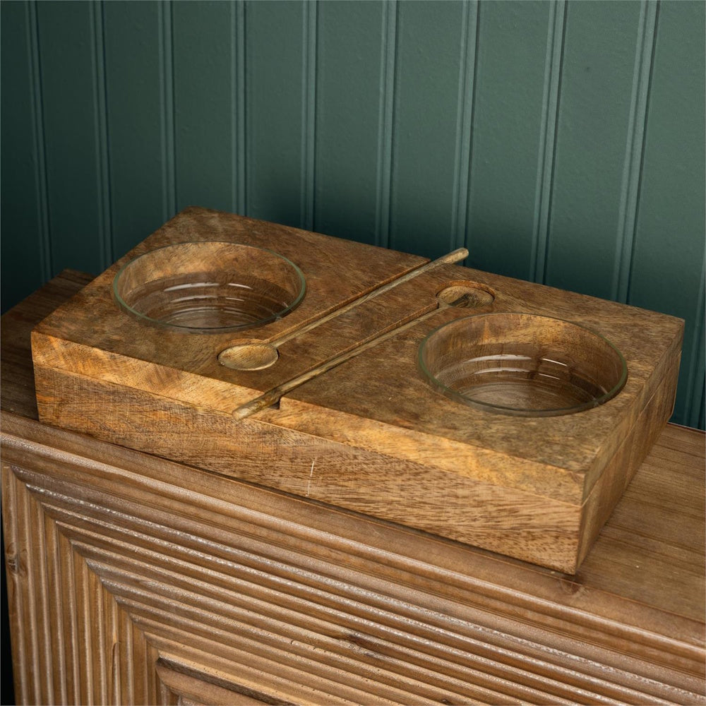 Wooden serving board with two glass bowls and matching spoons resting on a wooden cabinet.