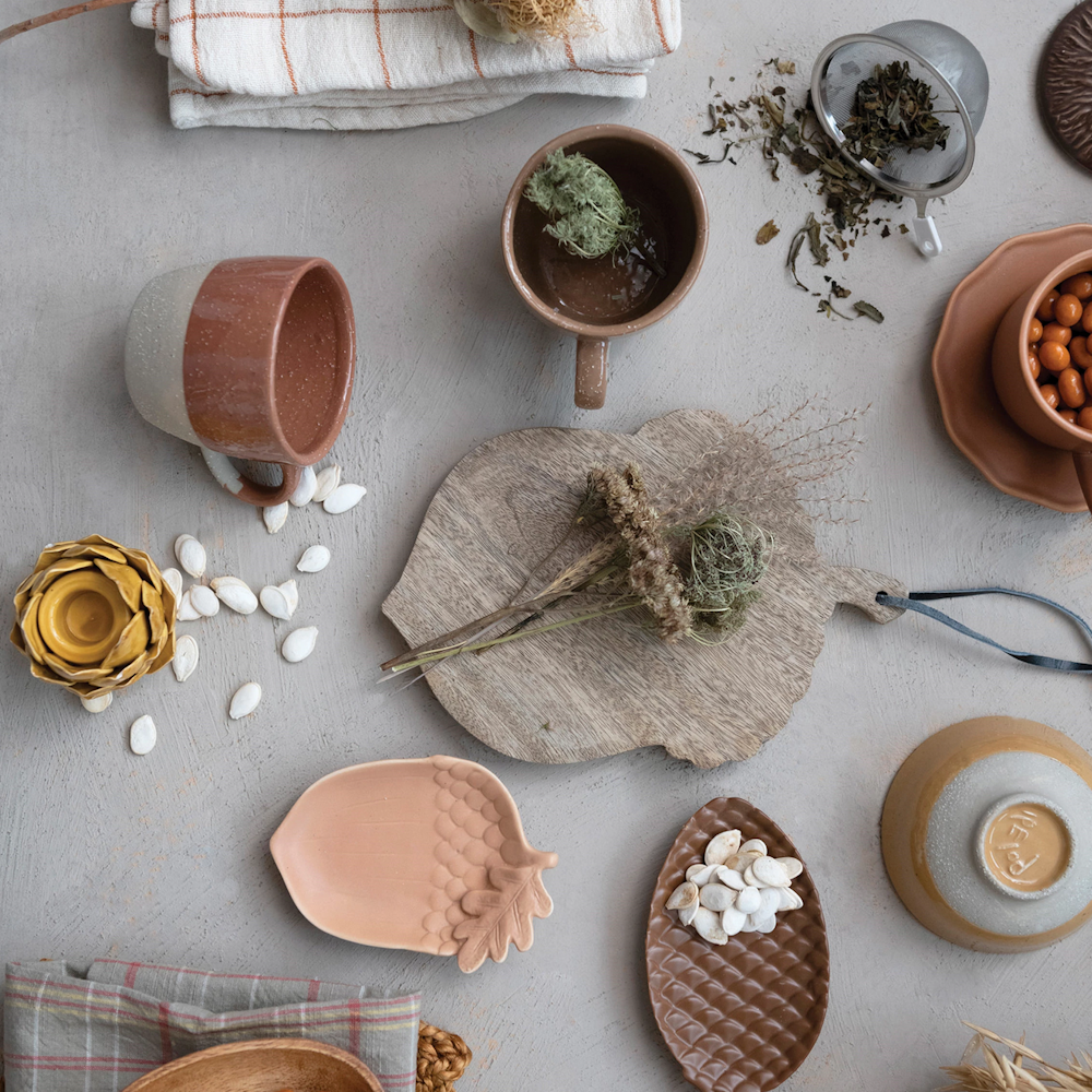 Top-down view of an acorn-shaped wooden cutting board surrounded by earthy ceramic mugs and dishes.