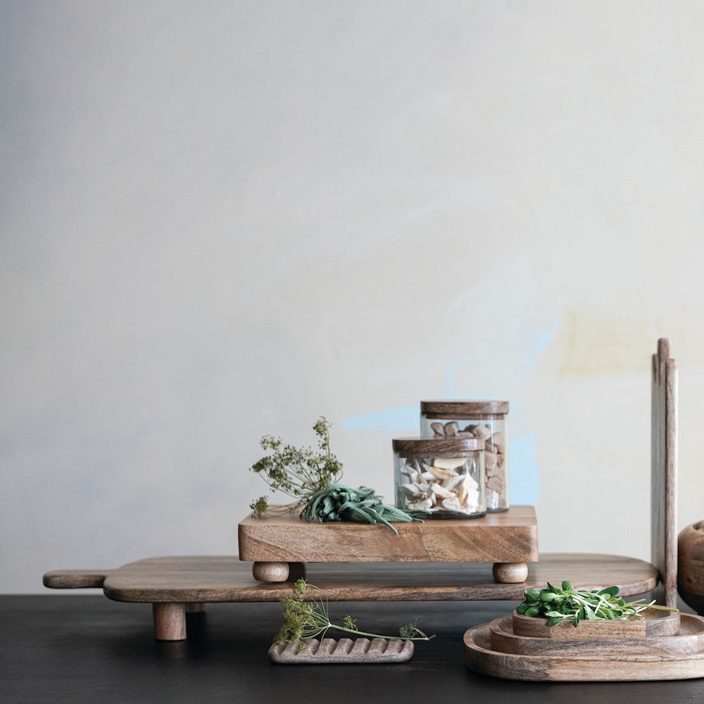 Footed mango wood cheese serving board holding glass jars and herbs, styled on a tabletop with other wooden boards.