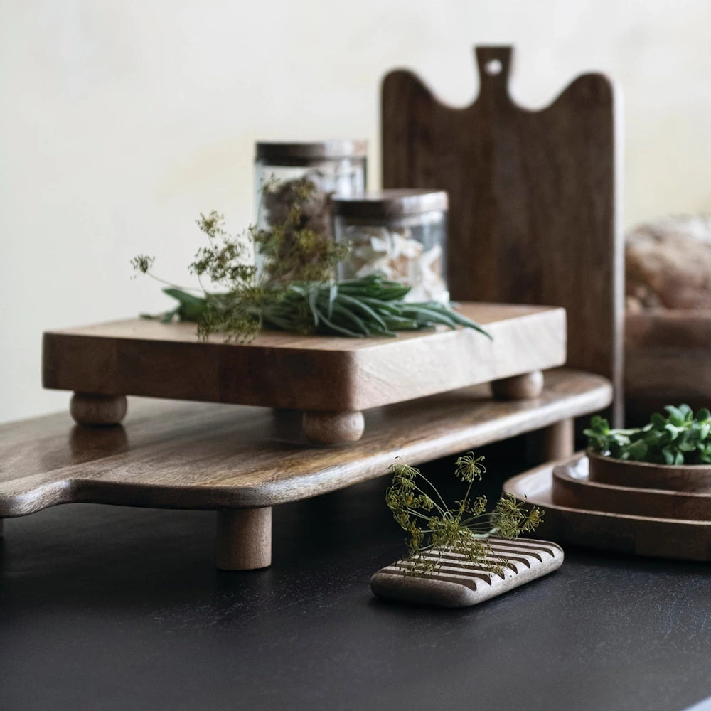 Mango wood footed cheese serving board with handle and tiered wooden kitchenware on a dark surface.