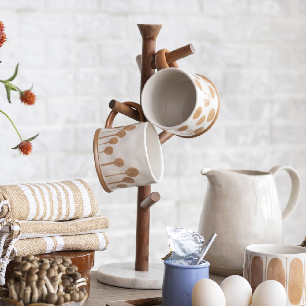 Wooden mug tree holding patterned cream mugs on a kitchen counter with eggs, a pitcher, and linens.