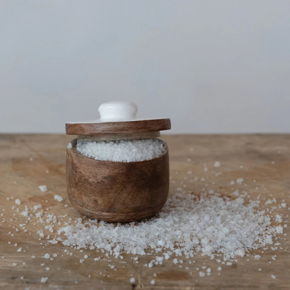 Coarse sea salt in a small wooden cellar with a white lid, scattered on a rustic wooden surface.