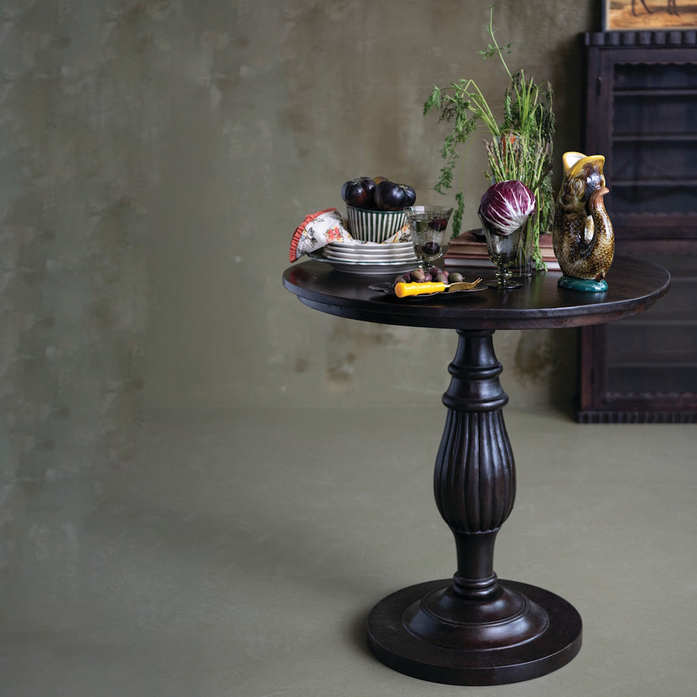 Round dark mango wood pedestal table styled with kitchenware and fresh produce.