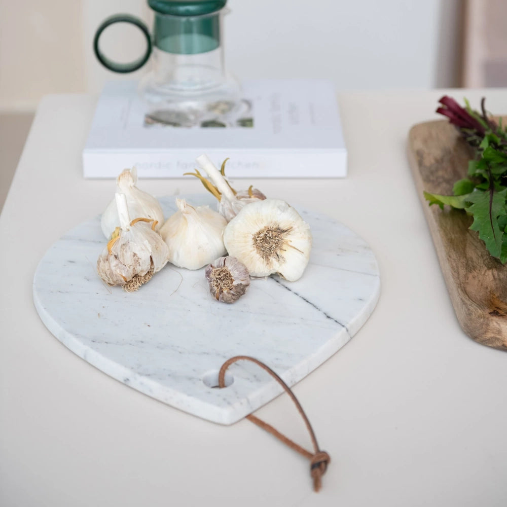 White marble cheese board with leather tie featuring garlic bulbs on a kitchen counter with a book and wooden board.