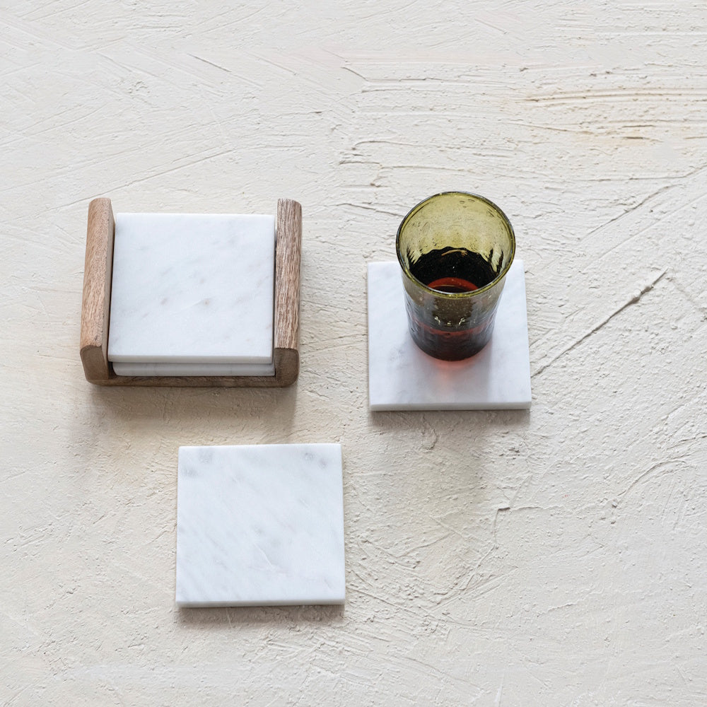 White marble square coasters set with a mango wood holder and green glass on a textured beige surface for home decor.
