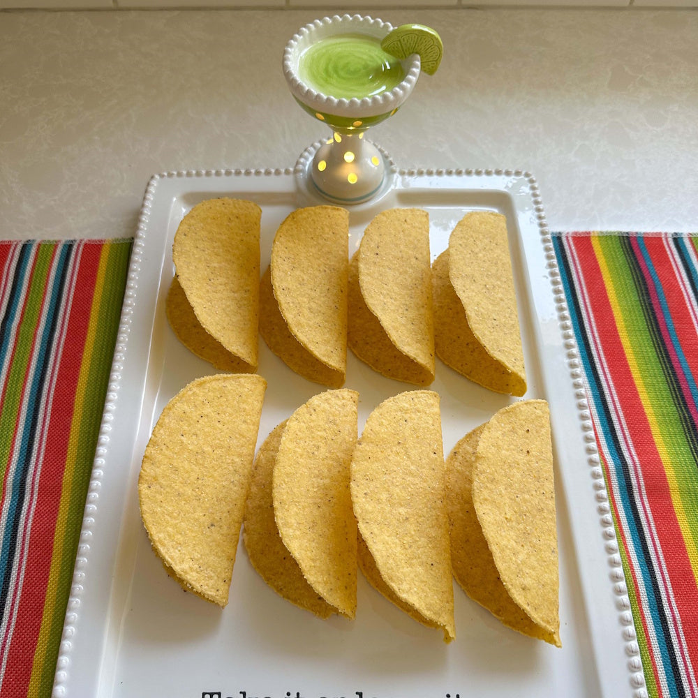 Hard taco shells arranged in two rows on a white platter with a decorative margarita-shaped dip bowl and a lime wedge.