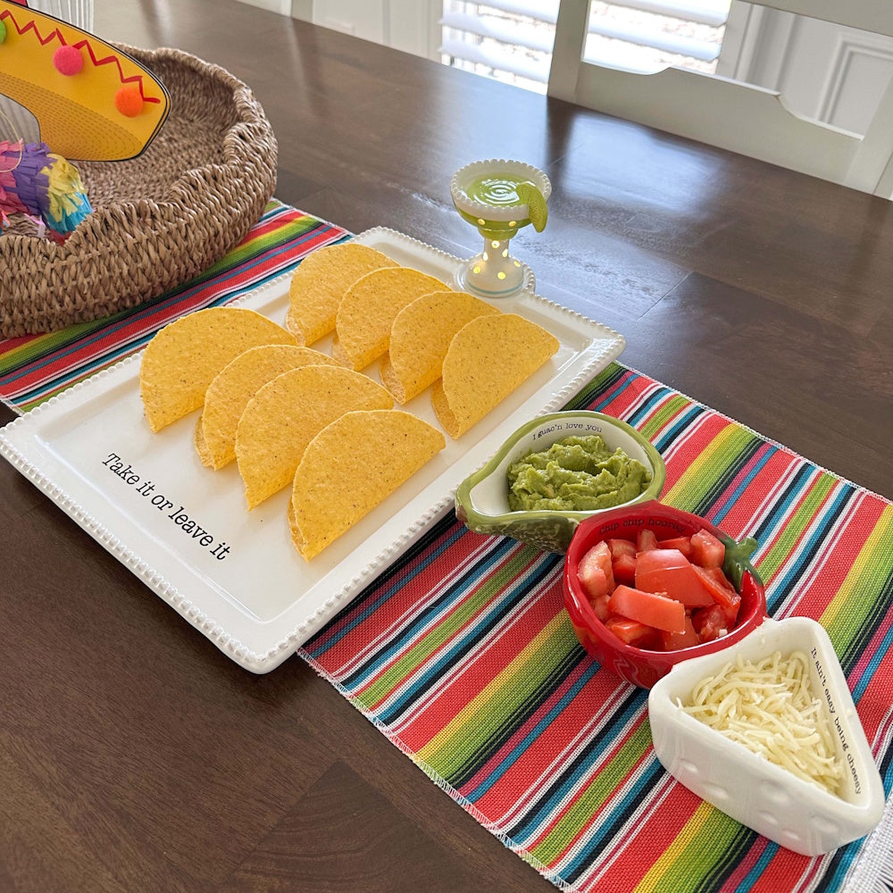 Taco shell tray with margarita light-up sitter, guacamole, tomatoes, and cheese on a colorful Serape table runner.