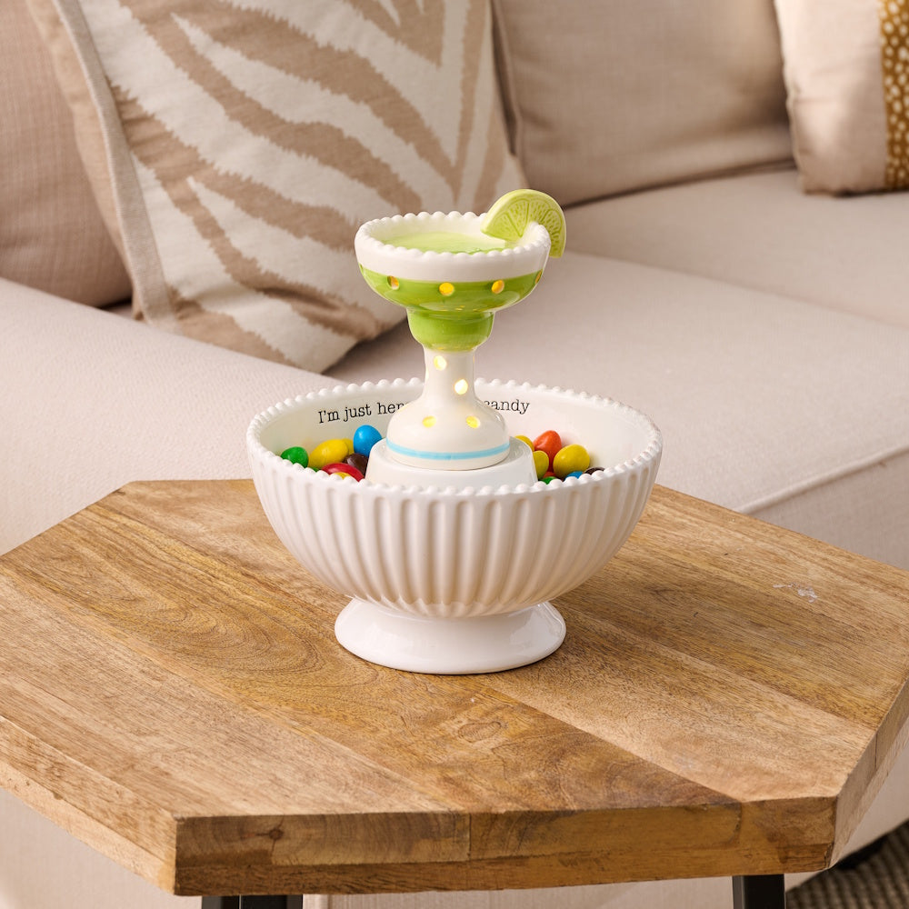 Light-up ceramic margarita sitter in a white candy bowl filled with colorful chocolates on a wooden side table.
