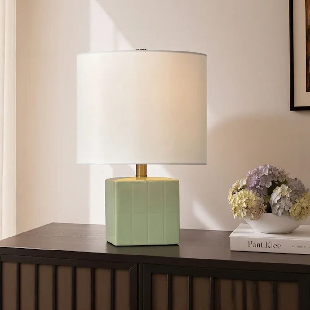 Mint green square ceramic lamp with a white drum shade on a dark wood sideboard next to a bowl of hydrangeas.