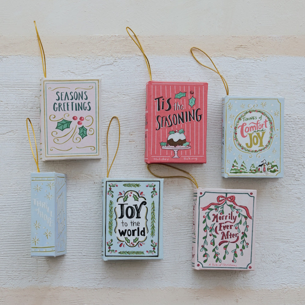 Set of six colorful miniature book Christmas ornaments with hanging gold strings on textured background.