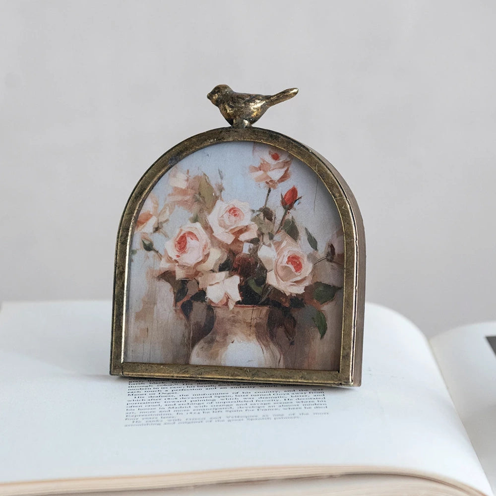 Arched gold metal frame with bird finial, featuring a vintage rose painting on an open book.