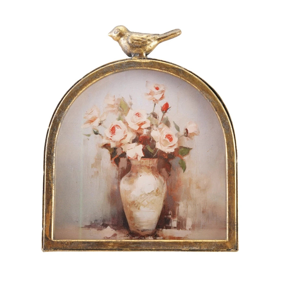 A framed floral print featuring pink roses in a vase with a small gold bird figurine on top.