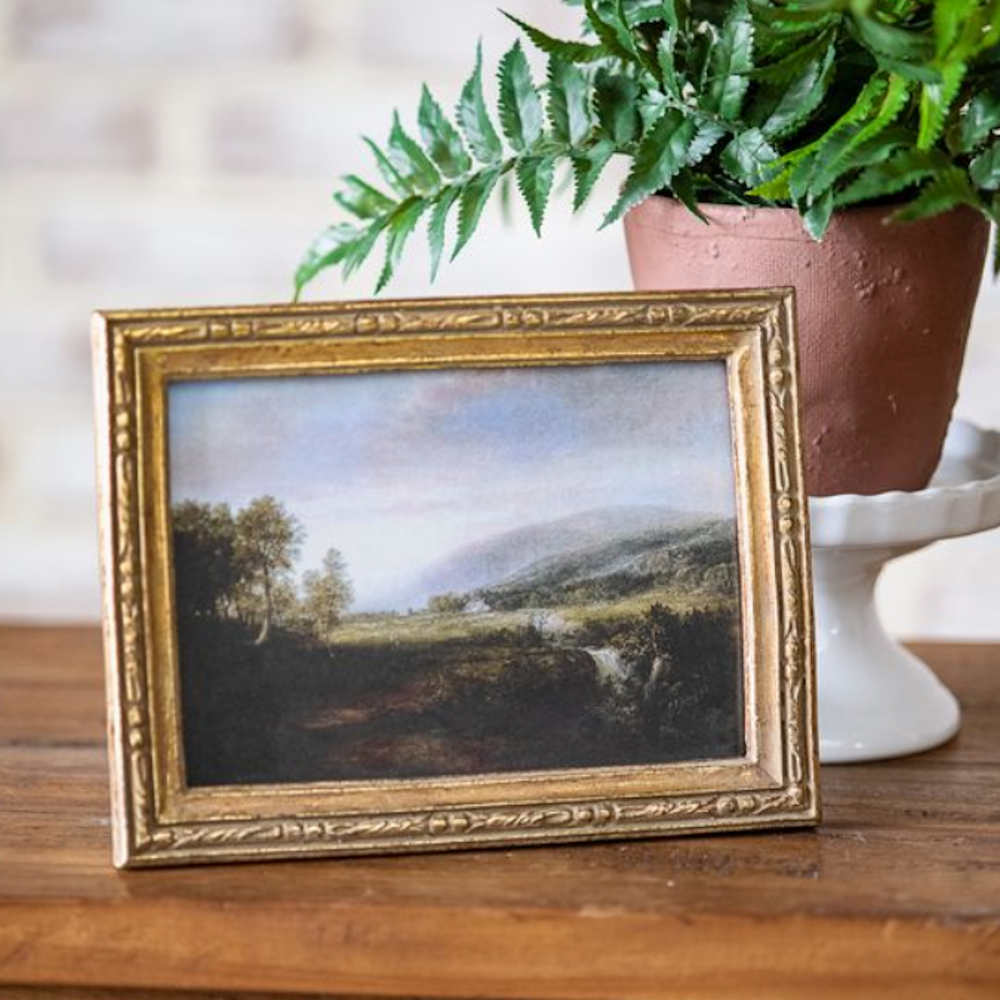 Framed countryside landscape painting in a triple-edge antique gold frame on a wooden surface.