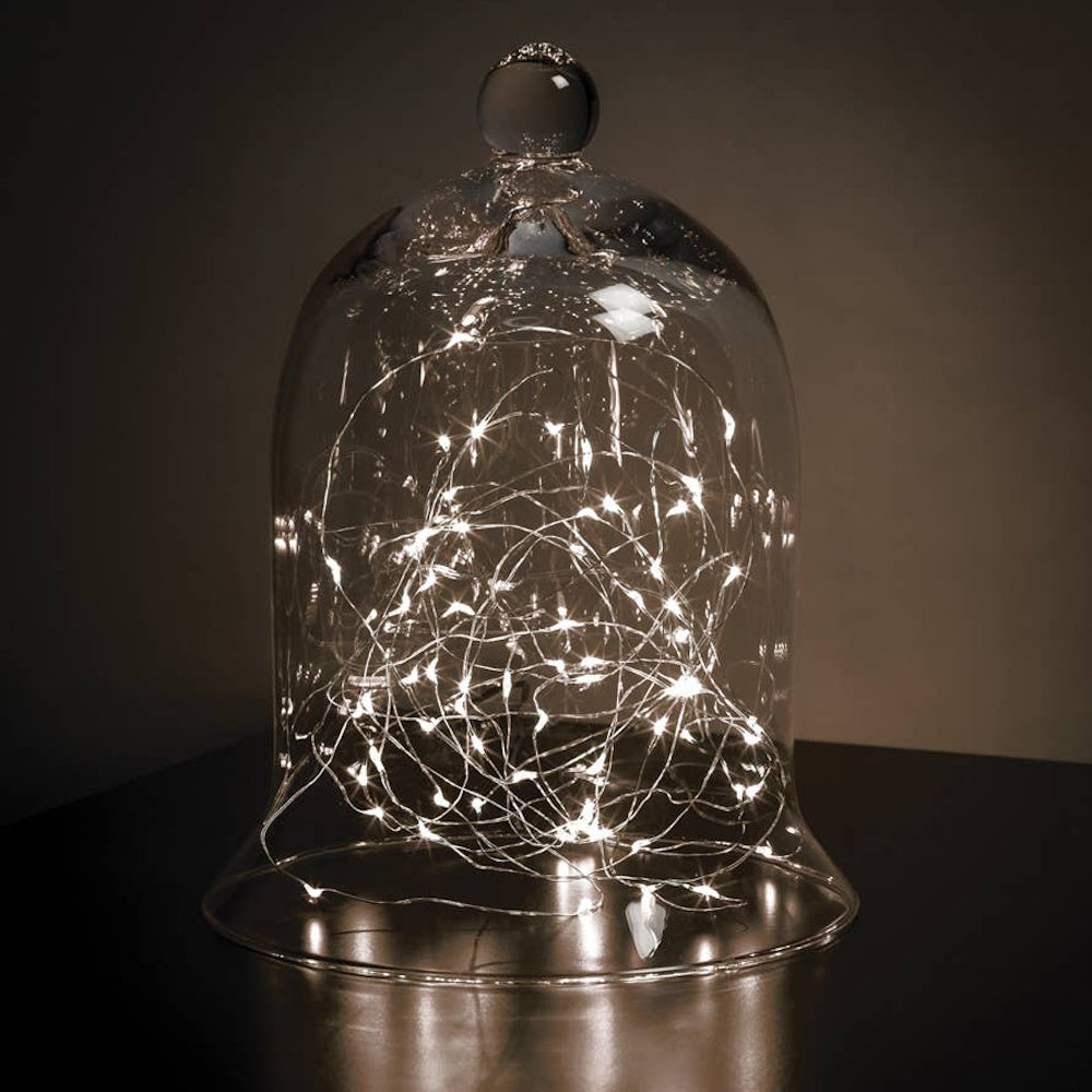 Clear glass bell-shaped cloche filled with glowing warm white fairy lights on a dark surface.