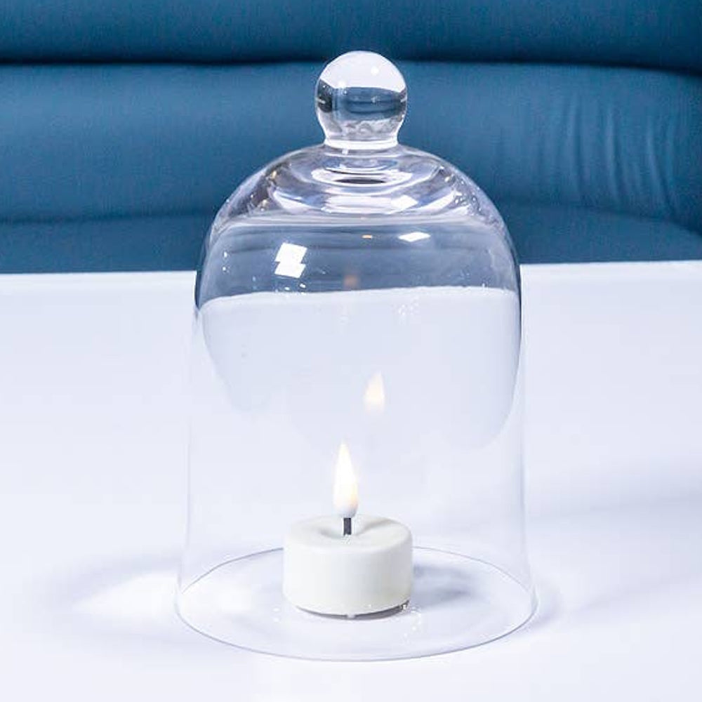 Clear bell-shaped glass cloche covering a lit white tea light candle on a table.