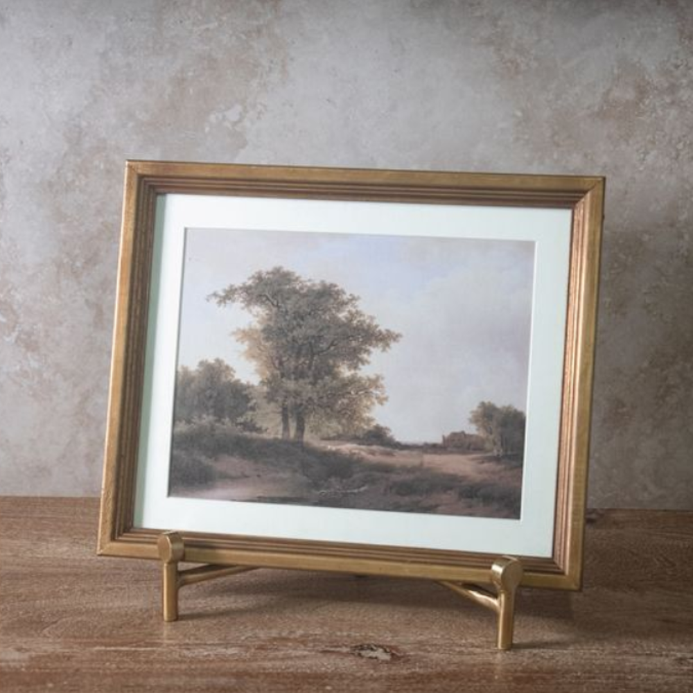 Framed landscape painting in a gold frame with a white mat, displayed on a gold easel.